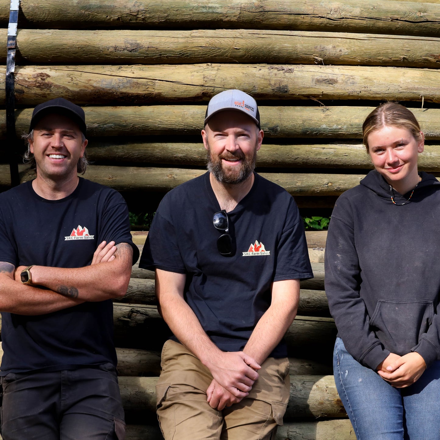 Three people posing in front of stacked fencing posts with a visible logo on their shirts.